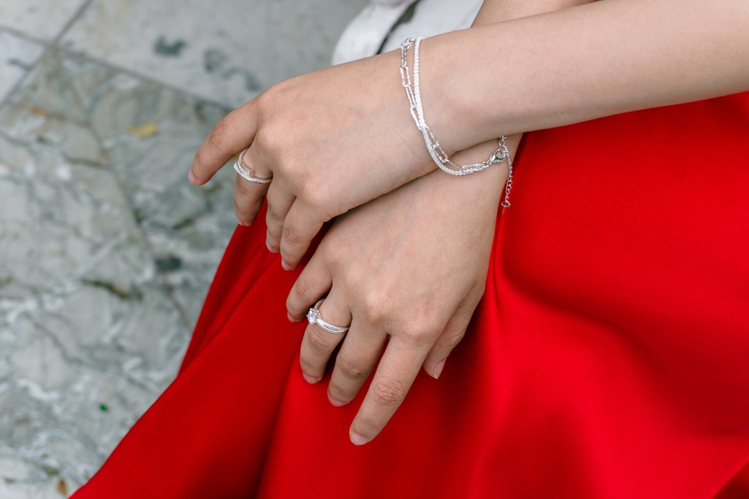 Two hands with rings and bracelets resting on red fabric.